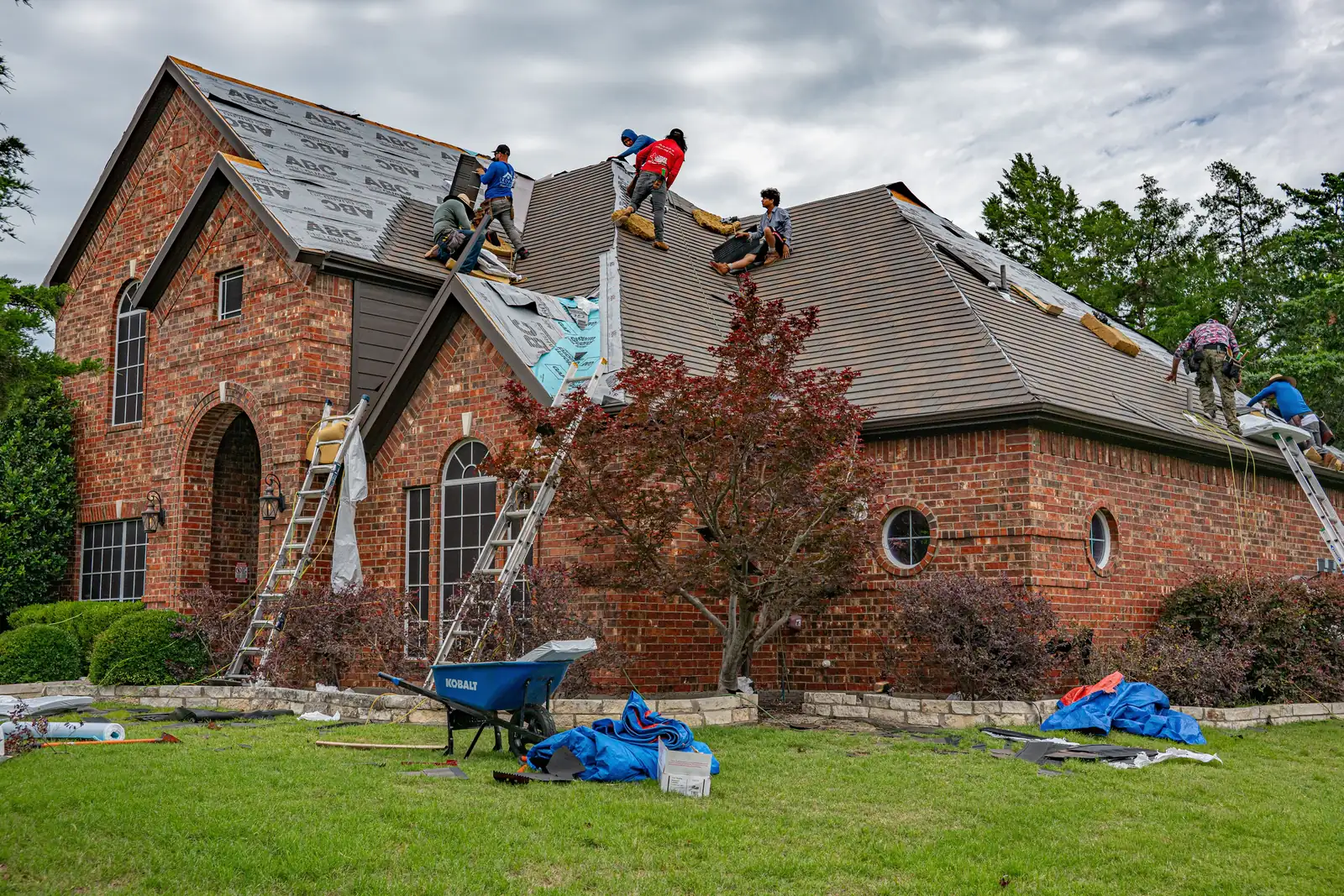 Roofing crew replacing the shingle roof on a two-story brick house, ladders against the walls