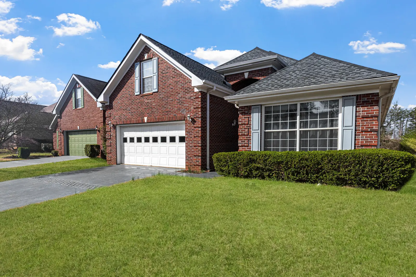 Red brick suburban home with a gabled gray shingle roof, two-car garage and green lawn